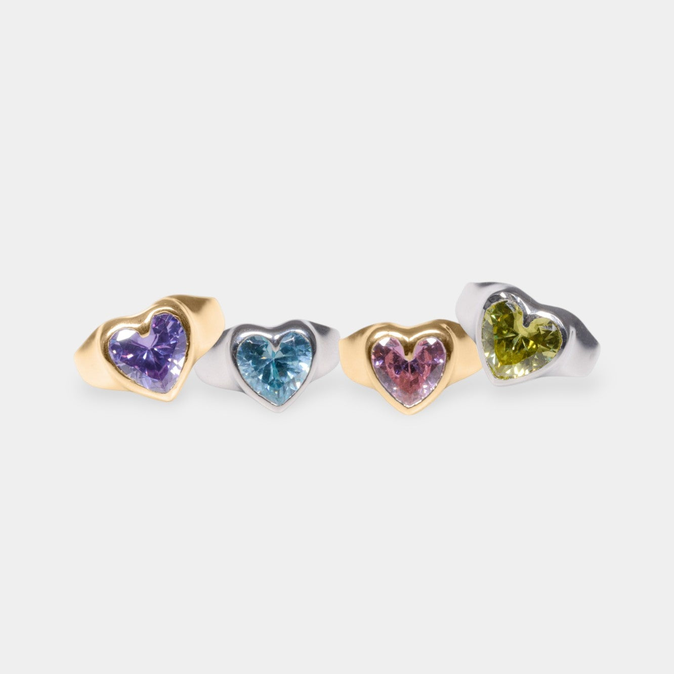 A collection of gold and silver rings with heart-shaped CZ stones in different colors: blue, pink, yellow, and purple, set against a white background.